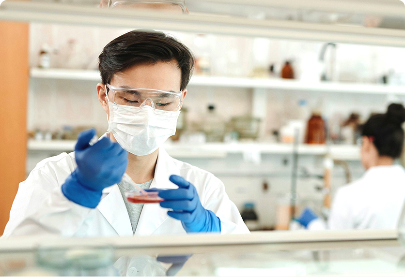 身着白大褂的科研人员戴护目镜、口罩与蓝手套，在实验室操作培养皿做实验，整体风格专业严谨，传递出生物制药领域专注研发的氛围。